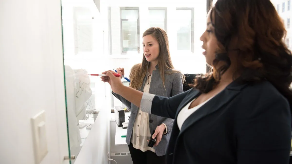 Two women in business attire write on a whiteboard.