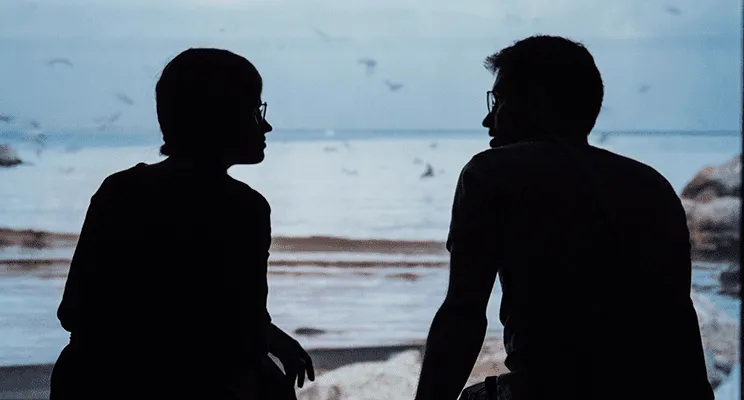 Two people turn to face each other while sitting silhouetted on a beach